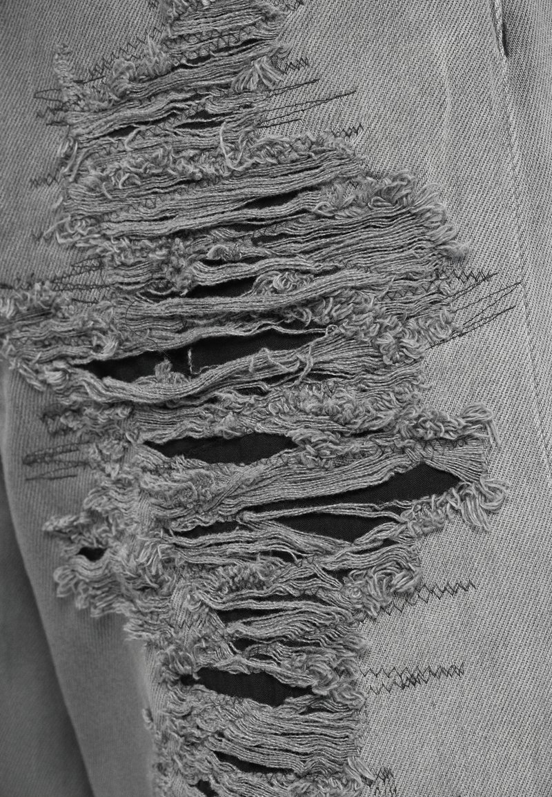 Grå denim med framträdande slitna fläckar som visar fransiga kanter och hål, med kontrasterande svart tyg under.