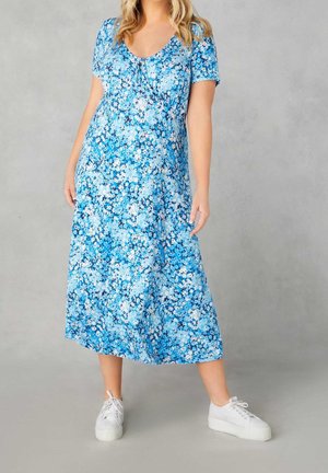 Frau trägt ein blaues Midikleid mit floralem Muster, kurzen Ärmeln und weißen Plateausneakern, steht vor einem grauen Hintergrund.