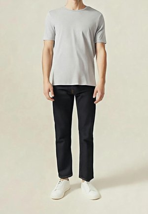 Homme portant un t-shirt gris clair à manches courtes, pantalon noir droit et baskets blanches, debout sur un fond uni.