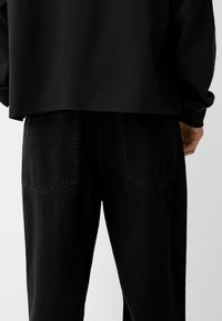 Črne jeans hlače s ravnim krojem, ki imajo dva zadnja žepa in rahlo teksturirano površino. V kombinaciji s črnim, skrajšanim zgornjim delom z dolgimi rokavi.