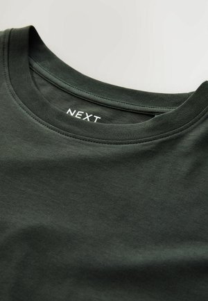 Camiseta de algodón verde oscuro con un escote redondo con ribete y una etiqueta blanca visible marcada "NEXT" en el collar interior.