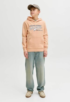 Persikanvärinen huppari graafisella tekstillä ja kengurutaskulla; yhdistetty vaaleansinisiin, väljämallisiin farkkuihin ja beigenvärisiin kenkiin. Yksinkertainen lippalakki täydentää asun.