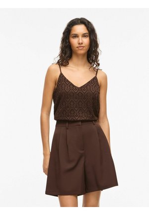 Göndör hajú nő ujjatlan barna mintás felsőt és magasderekú, barna, szabott rövidnadrágot visel, fehér háttér előtt.