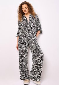 Donna con capelli ricci che indossa una camicia a maniche lunghe con stampa zebrina bianca e nera e pantaloni a gamba larga abbinati, insieme a sneakers beige.
