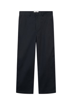 Person med langt hår står med fronten fremad, hænderne i lommerne, iført en hvid t-shirt med grafik, sorte bukser og hvide sneakers, på hvid baggrund.