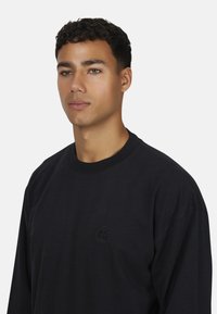 Jonge man met kort krullend haar draagt een eenvoudige zwarte lange mouwen shirt met een subtiele geborduurde logo op de borst tegen een witte achtergrond.