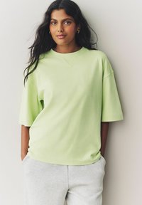 T-shirt en coton vert clair, surdimensionnée, à manches courtes et col rond, associée à un pantalon de survêtement gris, sur fond neutre.