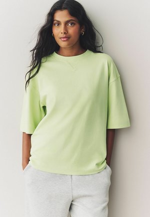 Lichtgroen oversized katoenen t-shirt met korte mouwen en een ronde hals, gecombineerd met grijze sweatpants, geplaatst tegen een neutrale achtergrond.