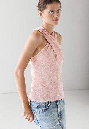 Femme portant un haut sans manches rose pâle à col halter avec des bretelles croisées et un jean bleu clair sur un fond uni gris clair.
