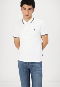 White short-sleeve polo shirt with a zip collar, black accent stripes, and a small embroidered design on the chest. Worn with blue jeans.
