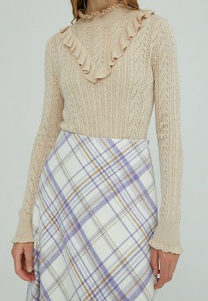 Pull beige en laine avec un col volanté et un motif texturé, associé à une jupe blanche à carreaux avec des lignes violet clair et marron.