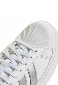 Witte sneaker met een bovenwerk van getextureerd leer, witte veters en metallic zilveren accenten aan de zijkanten. Afgeronde neus met zachte details.