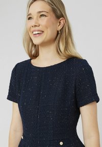 Robe bleu marine en tissu texturé, avec des manches bouffantes courtes, un col rond et des accents de boutons dorés à la taille.