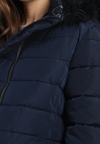 Marineblå puffjacka med horisontell sömnad, huva med fuskpäls och dragkedja fram. Slät textur med en blank finish.