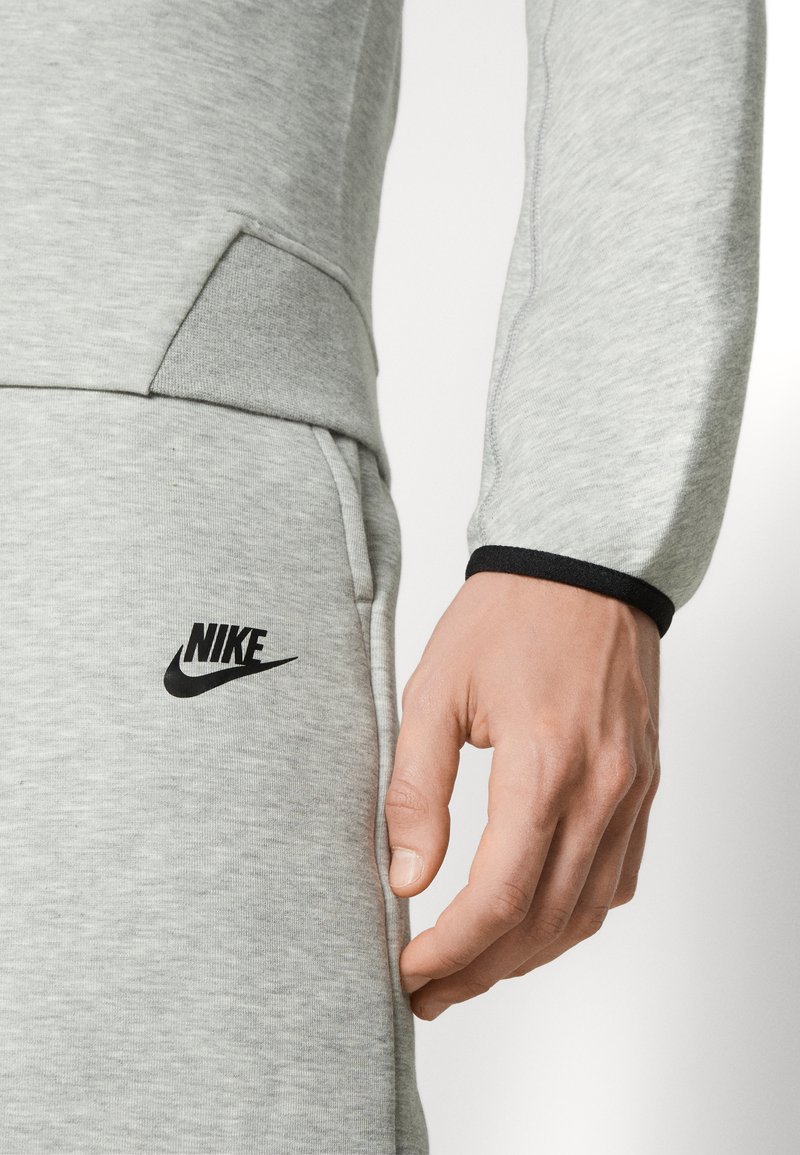 Sweat-shirt de sport gris à manches longues avec un logo noir sur la poitrine ; présente un poignet ajusté contrastant avec un design de poche.