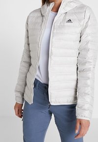 Jasnoszara kurtka puchowa z kapturem, z pionowym pikowaniem, pełnym zamkiem błyskawicznym oraz małym czarnym logo Adidas na lewej piersi.