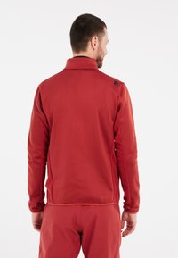 Rode softshell jas met een hoge kraag, getextureerde zijpanelen en een reflecterend logo op de schouder. Lichtgewicht, getailleerd ontwerp.