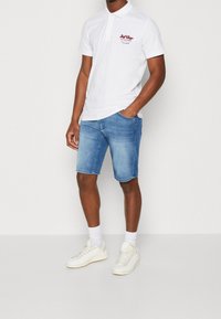 White short-sleeve polo shirt with embroidered detail, paired with light blue denim shorts and white sneakers, highlighting casual summer wear.