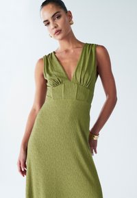 Groen mouwloze jurk met een diepe V-hals, gestructureerde stof en een gevormd lijfje, voorzien van een subtiel patroon en een elegante silhouet.