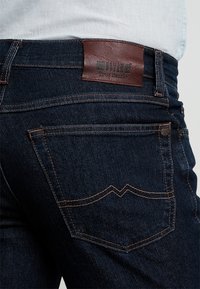 Tmavě modré džíny s přední kapsou a hnědým pro�šitím a hnědou koženou nášivkou s nápisem "Mustang True Denim" pod světlou košilí.