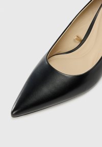 Zapato de tacón alto de cuero negro con punta afilada y una abertura en forma de V de bajo corte. Textura suave con un interior beige.