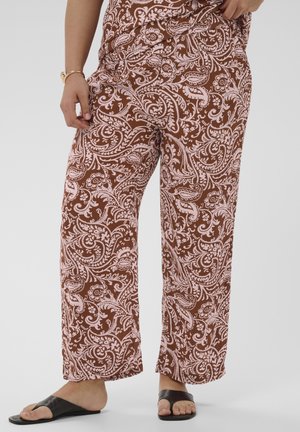 Mujer con pantalones marrones holgados con patrones florales y de paisley en rosa, y sandalias negras tipo chancla, de pie frente a un fondo liso.