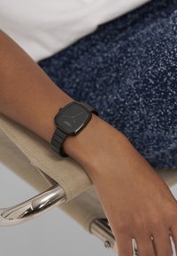 Sort rektangulært ur med mat finish og enkel urskive på en matchende rem, båret på et håndled hvilende på en sølvfarvet stol arm.