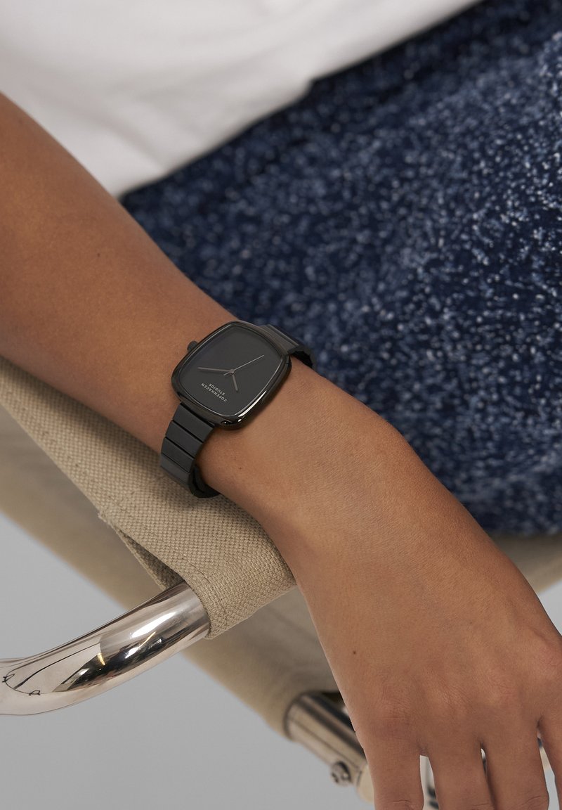 Sort rektangulært ur med mat finish og enkel urskive på en matchende rem, båret på et håndled hvilende på en sølvfarvet stol arm.