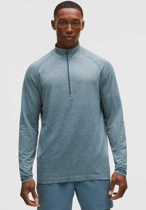 Mand iført en lyseblå langærmet sportstrøje med kvart lynlås og matchende blå shorts, stående mod en ensfarvet baggrund.