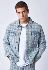 Veste en denim avec des tons bleu clair, motif en losanges, deux poches poitrine, devant à boutons et tissu texturé. Portée sur une chemise blanche.