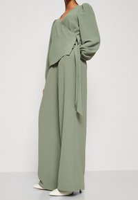 Grön, strukturerad jumpsuit med en omslagdesign, långa ärmar och knytband i midjan, kombinerad med vita klackskor med spetsig tå.