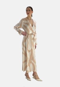 Robe longue beige avec des motifs crème abstraits, à enroulement et à manches longues. Associée à des chaussures beiges à bout pointu. Texture lisse.