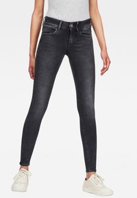 Jeans ajustados negros de mezclilla, con una cintura de altura media, bolsillos frontales y un sutil desgaste. Combinados con zapatillas blancas.