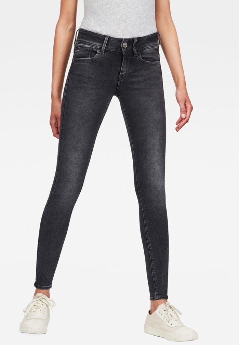 Jeans ajustados negros de mezclilla, con una cintura de altura media, bolsillos frontales y un sutil desgaste. Combinados con zapatillas blancas.