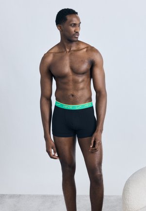 Μυώδης άνδρας με μαύρα μπόξερ εσώρουχα με πράσινη ζώνη, όρθιος ξυπόλυτος μπροστά σε απλό φωτεινό φόντο.