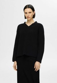 Maglione nero a maglia con scollo a V, caratterizzato da una texture a coste e maniche lunghe, indossato sopra una gonna nera.