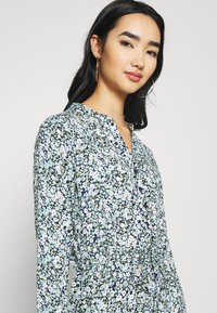 Abito button-up floreale con uno sfondo azzurro chiaro, piccoli fiori bianchi e verdi, maniche lunghe e vita cintata.