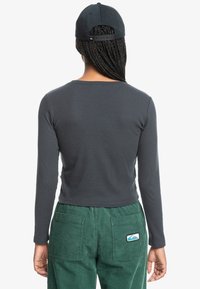 Gray long-sleeve shirt with a round neckline, paired with green corduroy pants featuring back pockets and a blue and white logo patch.