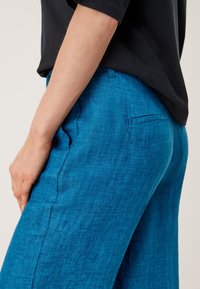 Gros plan d'une personne portant un pantalon en lin bleu vif avec une poche latérale et un haut noir à manches courtes, la main reposant près de la hanche.