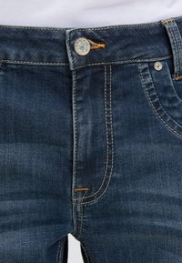 Dark blue denim jeans close-up showing button, zipper, pockets, and orange stitching details on a plain light background.