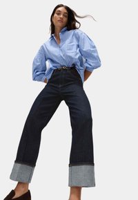 Donna che indossa una camicia button-up a righe blu con maniche a sbuffo, jeans larghi scuri con ampi risvolti e scarpe slip-on scure.