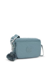 Light blue fabric crossbody bag with a round logo, zip closure, and soft furry keychain accent. Adjustable strap and rectangular shape.