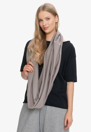 Młoda kobieta o długich blond włosach nosi luźny szary szalik infinity, czarną bluzkę z krótkim rękawem i jasnoszare spodnie na białym tle.