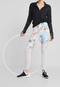 Osoba w czarnej bluzie z długim rękawem, legginsach w kwiatowy wzór i czarno-białych trampkach trzyma białą hula hoop.