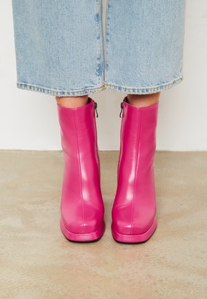 Femme portant des bottines en cuir rose vif avec des fermetures éclair, debout sur un sol beige, portant un pantalon en jean bleu clair raccourci.