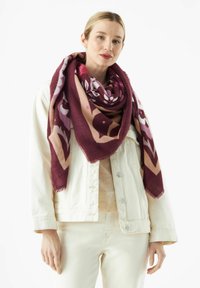 Bordeauxrode en crèmekleurige oversized sjaal met een geometrisch patroon, zachte textuur en franjes aan de randen, gedragen over een lichte trui en jas.