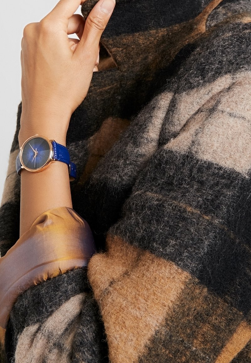 Gros plan d’une main ajustant un manteau en laine à carreaux, montrant une montre-bracelet à bracelet en cuir bleu et un poignet de chemise en satin marron brillant.