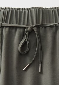 Close-up of olive green elastic waistband with tied drawstring and metal aglets on fabric pants.