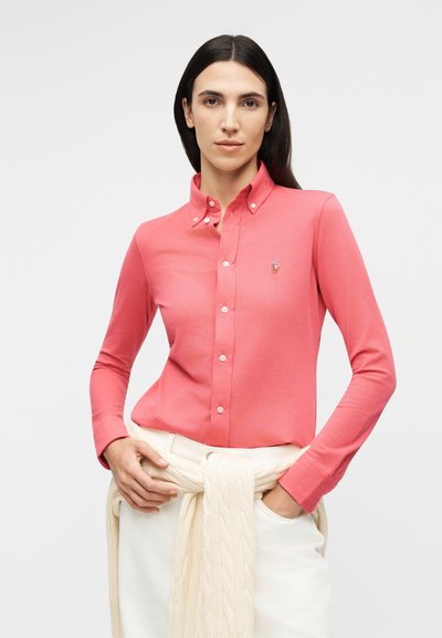 Nő korallrózsaszín, gombos ingben, apró hímzett logóval, krémszínű kötött pulóverrel a derekán kötve.
