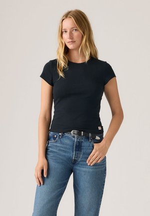 Žena s dlhými blond vlasmi, oblečená v priliehavom čiernom krátkoročnom tričku a modrých džínsoch s čiernym opaskom, stojaca pred jednoduchým pozadím.